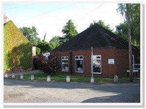Community Library & Coffee Shop A small brick building with a sloping roof, windows, and signs, beside a leafy street and a vine-covered wall.