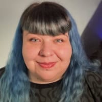 Smiling person with blue wavy hair, fringe, and a nose ring, wearing a dark patterned top.