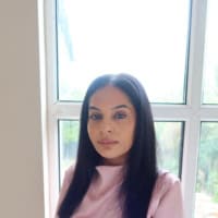 Woman with long black hair in a light pink top standing in front of a bright window.